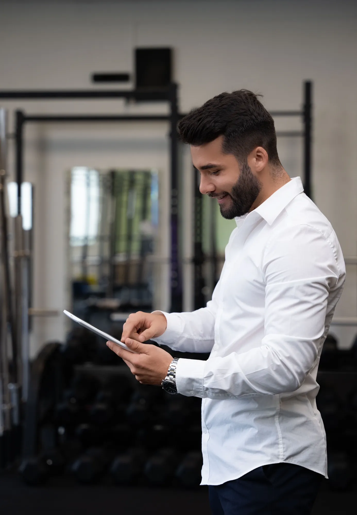 Jeune homme barbu en chemise blanche consultant une tablette devant des équipements de musculation