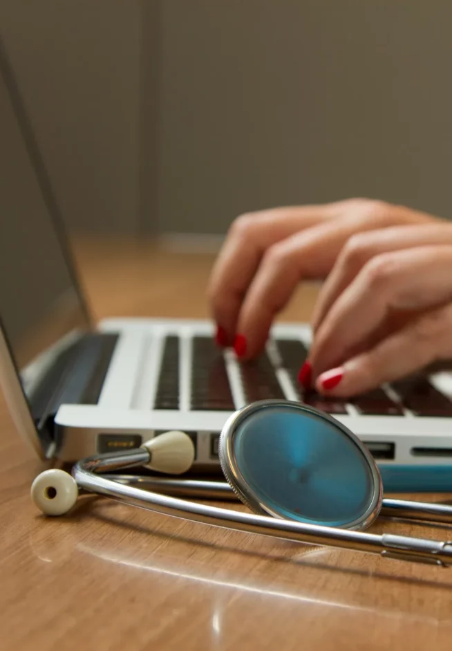 Professionnelle de santé saisissant des données sur un ordinateur portable, stéthoscope posé sur le bureau devant l’écran.